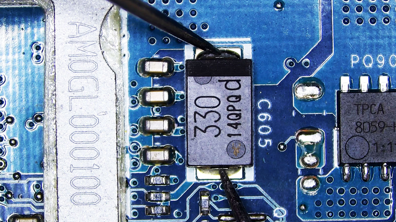 Close-up of a computer circuit board with capacitor C605 marked 330 being measured at both ends with multimeter leads.