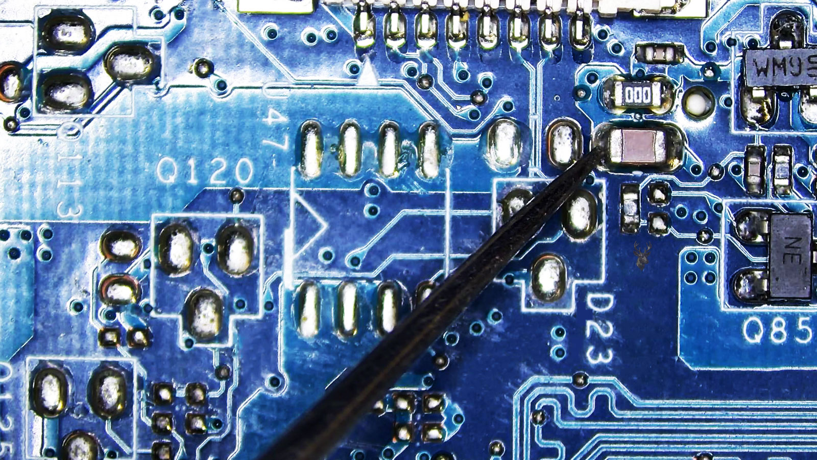 Close-up of a computer circuit board with a rear multi-pin connector, transistors, fuse, capacitors, and a multimeter probe testing an SMD capacitor near the fuse.