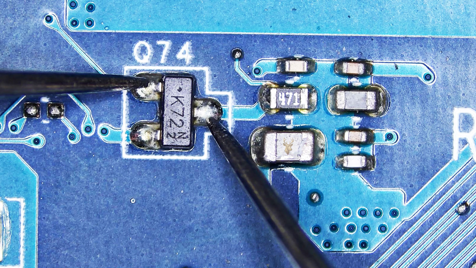 Close-up of a computer circuit board with capacitors, coils, a fuse, and a K72N2 transistor labeled Q74 being measured with a multimeter.