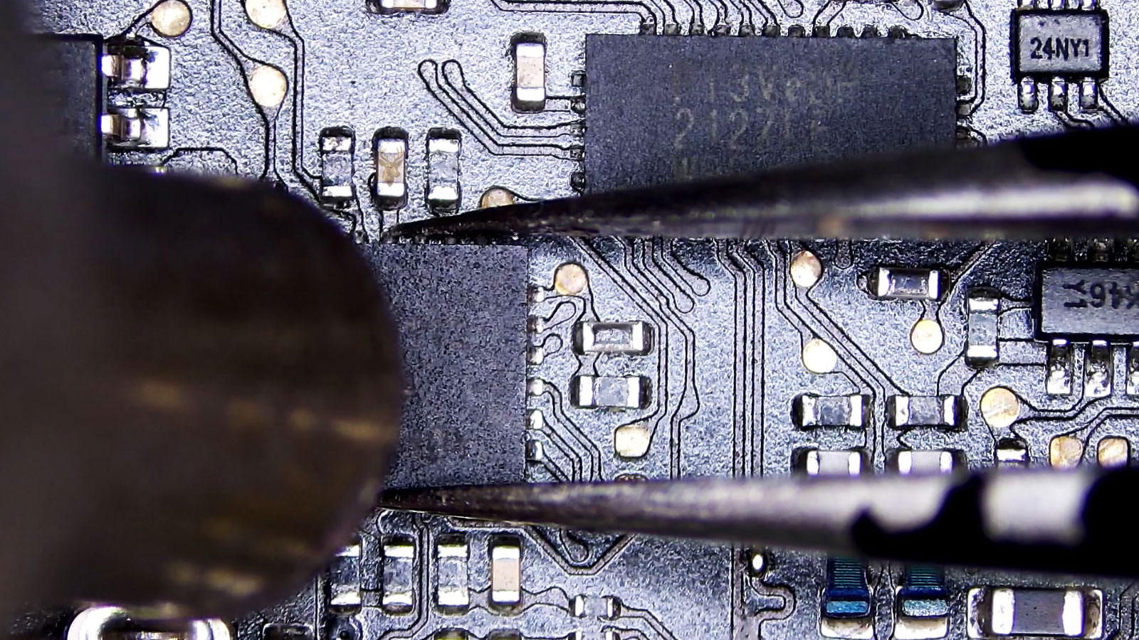 Close-up of a computer circuit board with tweezers holding a central IC chip while a hot air nozzle points toward it.