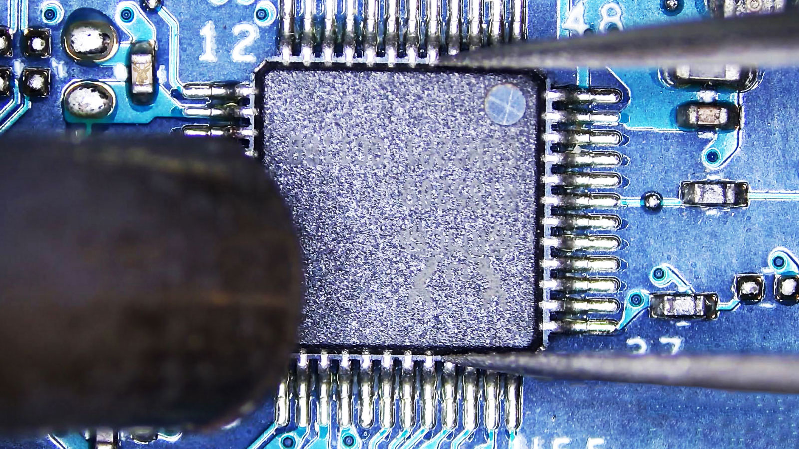 Close-up of a computer circuit section with a 48-pin IC chip, where precision tweezers and a hot air rework station nozzle are positioned to remove the chip from the circuit board.