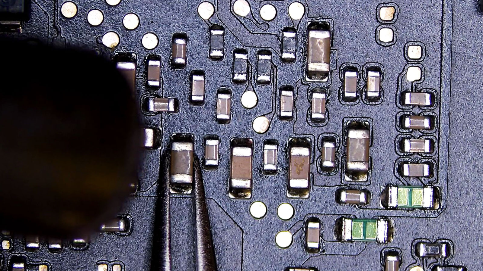 Tweezers holding a capacitor while a hot air tool heats it on a computer circuit board with surrounding electronic components.