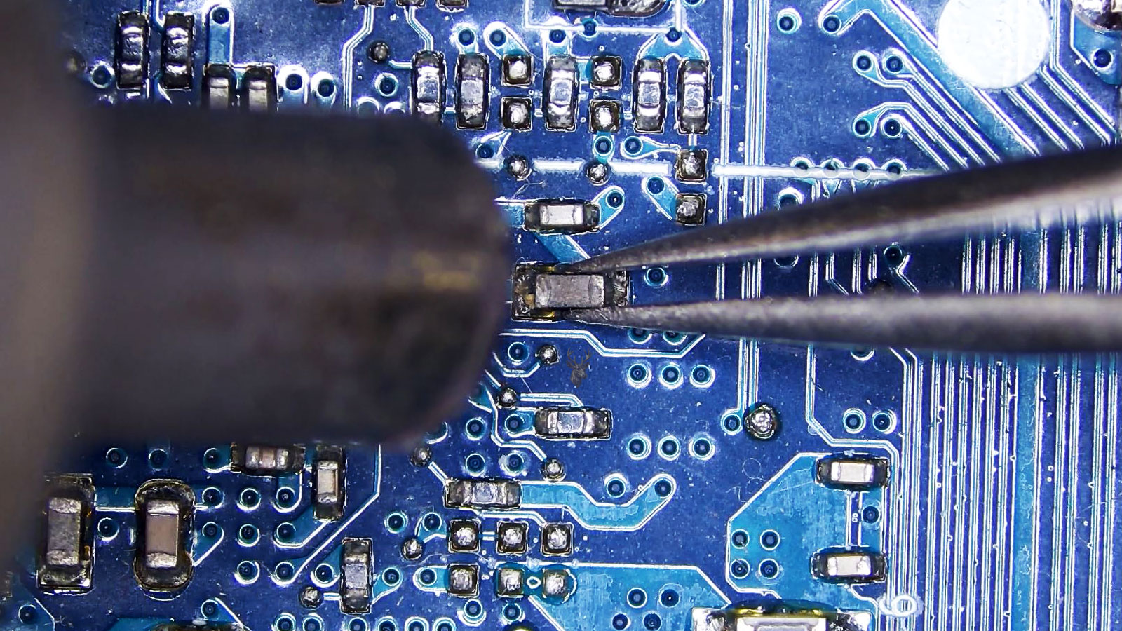 Tweezers holding a darkened SMD capacitor while a hot air rework nozzle heats it on a computer circuit board.