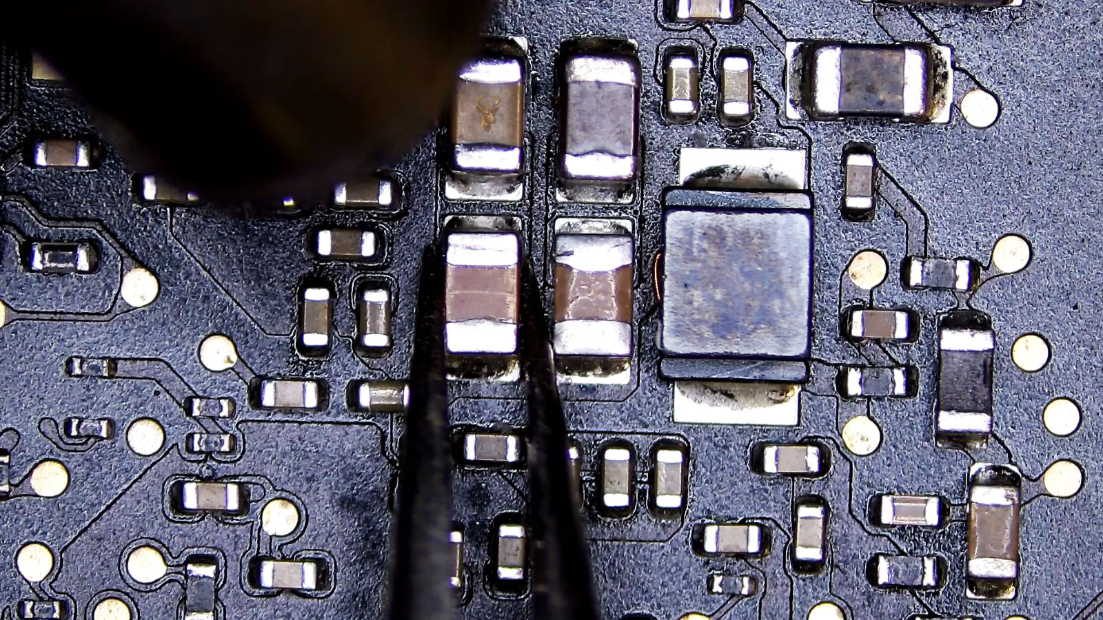 Tweezers holding the cracked SMD capacitor while a hot air gun nozzle points toward it.