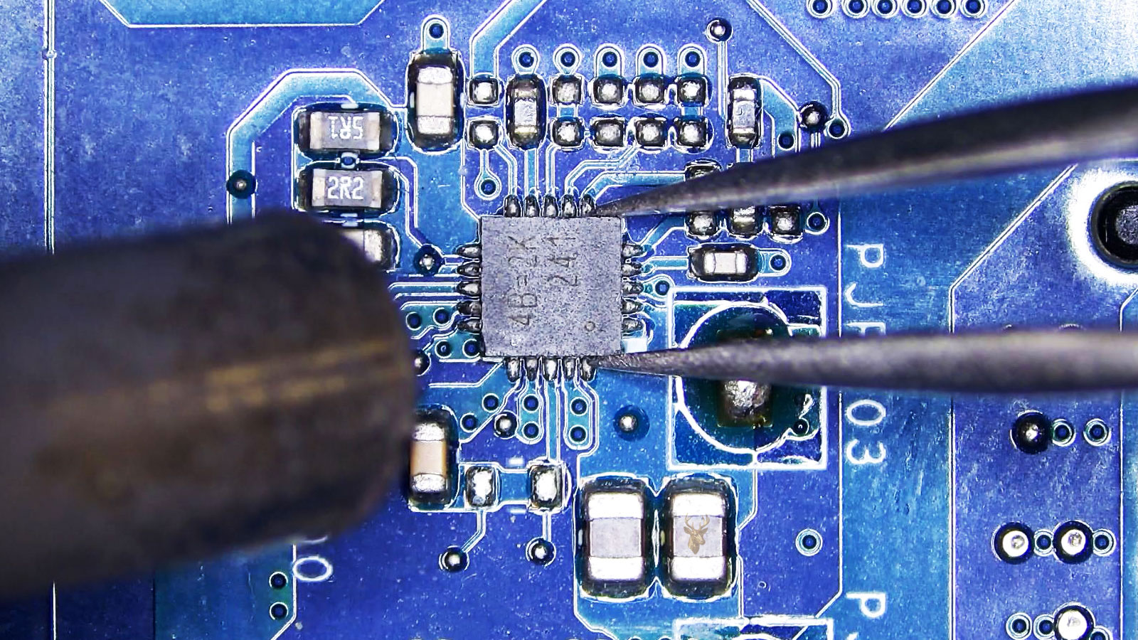 IC controller held by tweezers among capacitors, resistors, and coils while a hot air station prepares to remove it.
