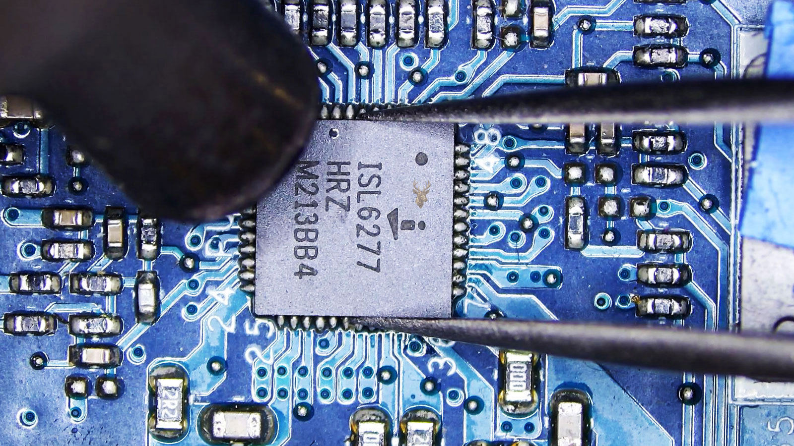 Tweezers holding the ISL6277 IC while a hot air station nozzle points at it in a densely populated circuit section.