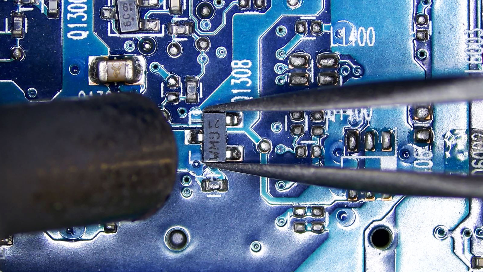 Q1308 transistor held by tweezers while a hot air station nozzle points at it near surrounding transistors, capacitors, and resistors.