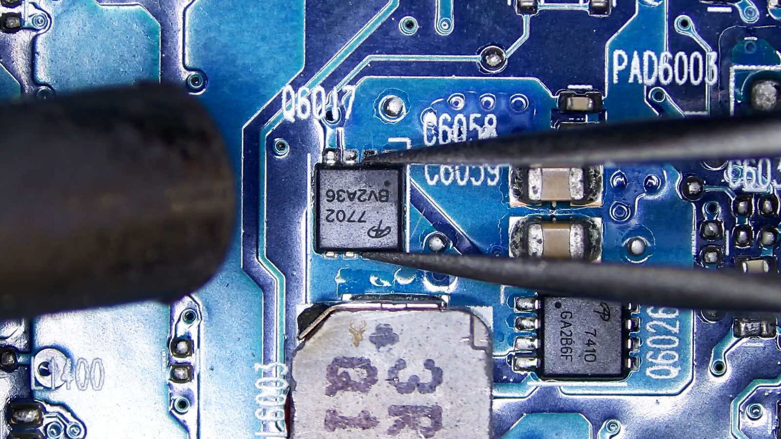 Q6017 MOSFET held by tweezers while a hot air station nozzle points at it near capacitors, coils, and another MOSFET.