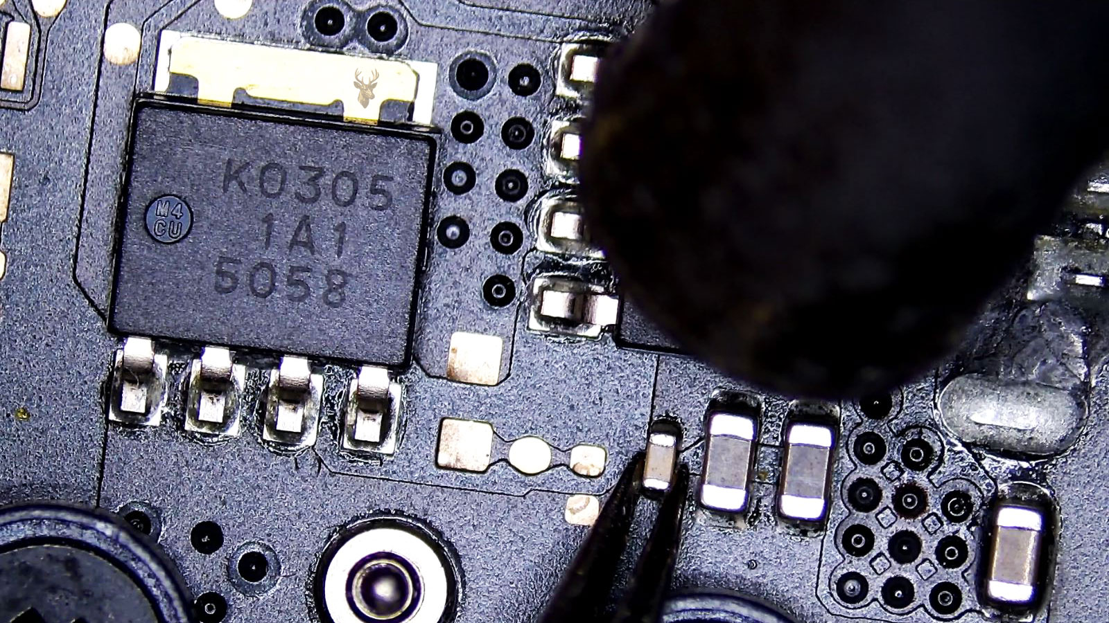 Small capacitor held by tweezers beneath two MOSFETs while a hot air station nozzle points at it.